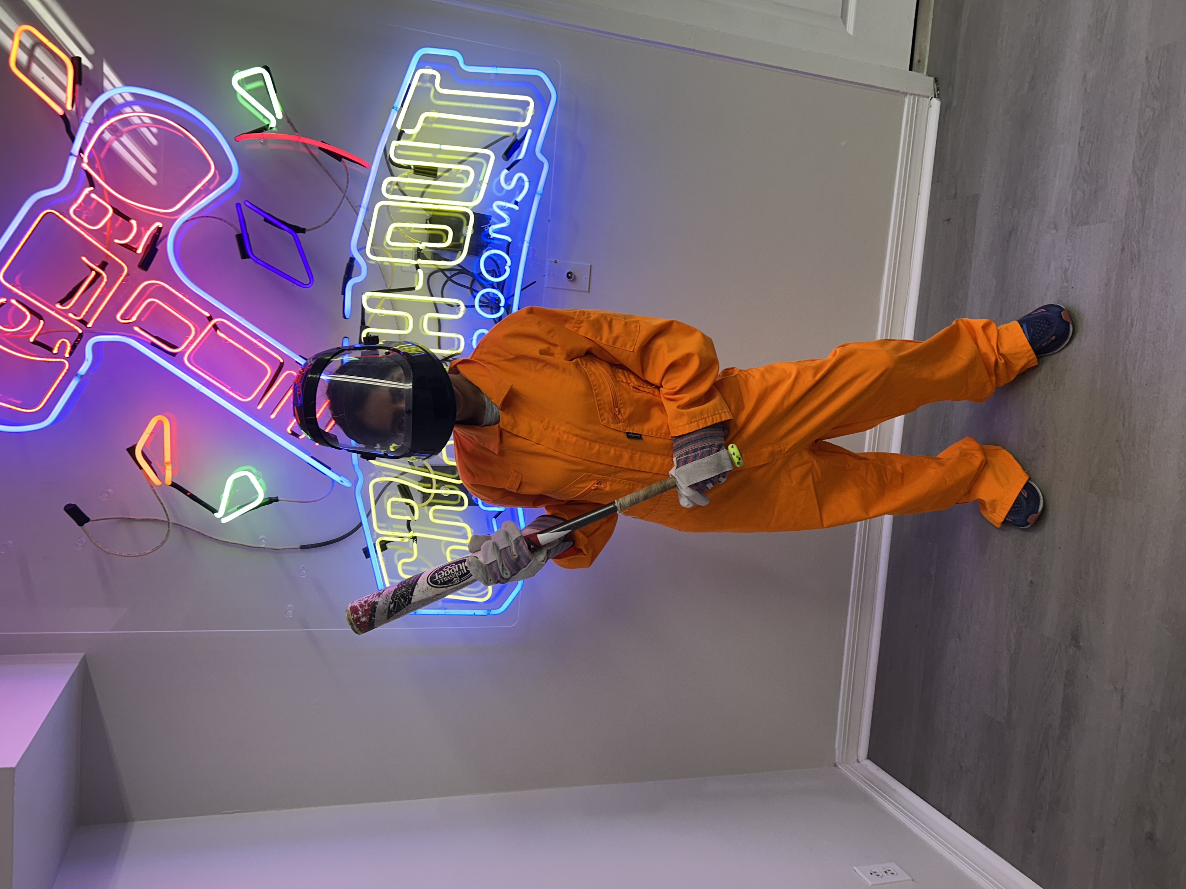 Crash Out customer in orange jumpsuit posing with bat in front of Crash Out neon sign