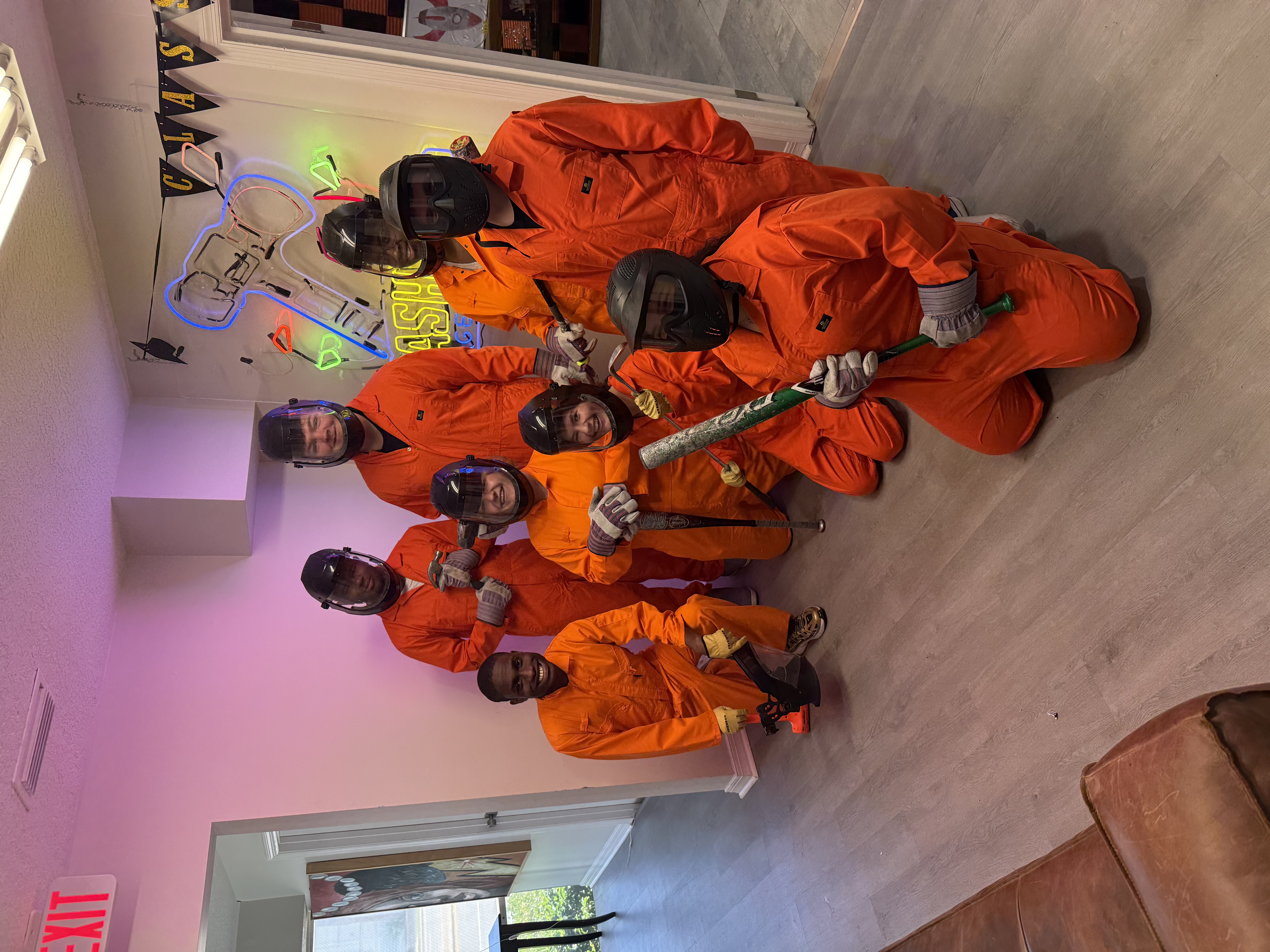 Eight Crash Out guests in orange jumpsuits holding smash tools and posing together